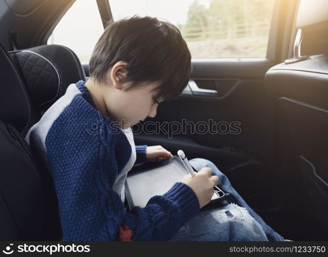 Happy young boy using a tablet computer while sitting in the back passenger seat of a car with a safety belt, Child boy drawing on smart pad,Portrait of toddler entertaining him self on a road trip.