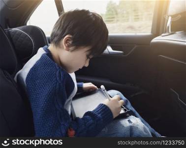 Happy young boy using a tablet computer while sitting in the back passenger seat of a car with a safety belt, Child boy drawing on smart pad,Portrait of toddler entertaining him self on a road trip.
