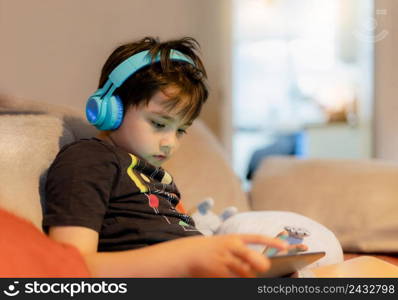 Happy young boy wearing headphone for playing game online on internet with friends, Kid sitting on sofa reading or watching cartoon on tablet Child relaxing at home in the morning on weekend