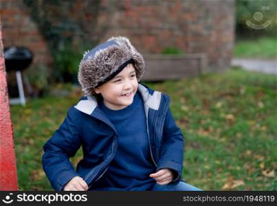 Happy young boy wearing warm clothes sitting in autumn park, Portrait cute boy having fun time playing outdoors in Fall season, Candid shot cheerful child looking out with smiling face.