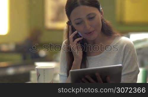 Happy young brunette enjoying her time in cafe. She having lively phone talk while using touch pad