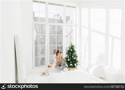 Happy young brunette woman and domestic animal decorate New Year tree at home. Cozy domestic interior in bedroom. Christmas firtree decoration. Winter holidays, people and celebration concept.