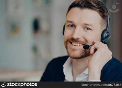 Happy young business professional or executive manager wearing suit and headphones with microphone smiling at camera while working from home or office, selective focus. Job and occupation concept. Smiling man in headset looking at camera with smile while having web conference