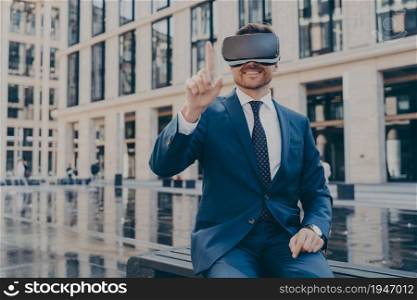 Happy young businessman in blue suit using virtual reality goggles while sitting on bench, controlling with his finger, excited man testing VR headset outdoors, office buildings in blurred background. Young businessman in blue suit using virtual reality goggles while sitting outdoors