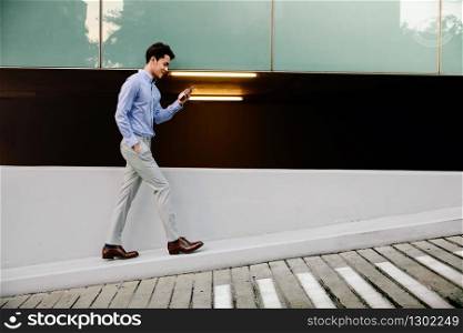 Happy Young Businessman in Casual wear Using Mobile Phone while Walking by the Urban Building Wall. Lifestyle of Modern People. Side View. Full Length
