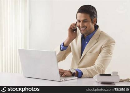 Happy young businessman on call while using laptop in office