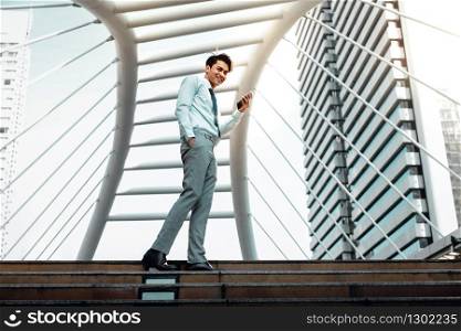 Happy Young Businessman Using Mobile Phone in the City. Lifestyle of Modern People. Low Angle View. Full Length. Looking at Camera and Smiling. Urban City as background
