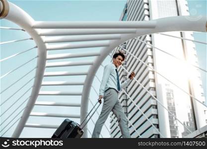 Happy Young Businessman Using Mobile Phone while Walking with Suitcase in the City. Lifestyle of Modern People. Low Angle View. Full Length. Modern Urban City as background