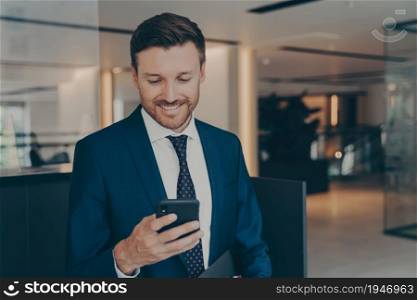 Happy young businessman using smart phone standing in office hallway, smiling male professional holding smartphone while reading sms from colleagues, enjoying corporate technology on mobile device. Happy young businessman using smart phone standing in office hallway