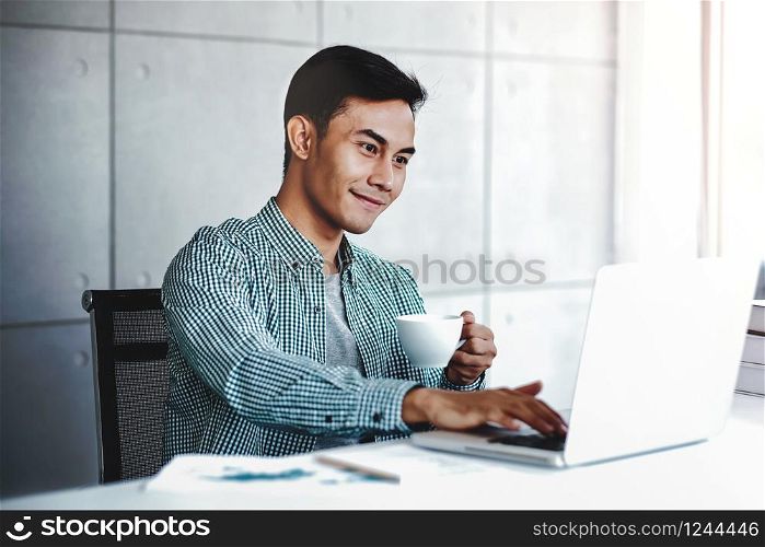 Happy Young Businessman Working on Computer Laptop while Drinking Coffee in Office