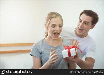 Happy young caucasian couple celebrating at home. Handsome boyfriend man gives his girlfriend with gift box surprisingly.