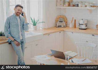 Happy young caucasian man holding cellphone while having conversation with client, standing in modern kitchen and enjoying good business talk while working distantly from home. Happy young caucasian man having conversation on cellphone in kitchen