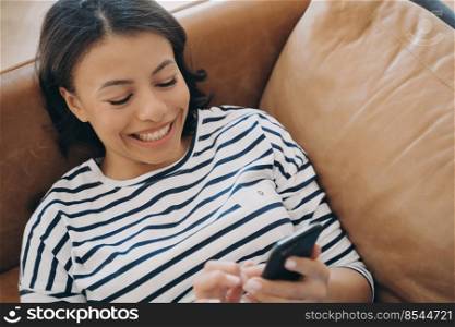 Happy young caucasian woman lying on leather couch. Gorgeous hispanic lady is texting on mobile phone and smiling, receiving and sending messages. Leisure and relaxation. Technology using at home.. Lady is texting on mobile phone and smiling. Happy young caucasian woman lying on leather couch.