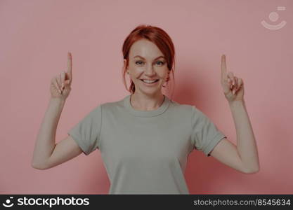 Happy young caucasian woman pointing upwards with index fingers and smiling cheerfully while promoting advertising product, isolated over pink background. Advertisement concept. Happy young caucasian woman pointing upwards with index fingers and smiling cheerfully
