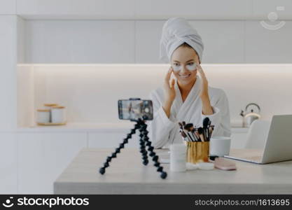 Happy young Caucasian woman records beauty blog on mobile phone standing on tripod goves advice how to use cosmetic patches shoots video wears bathrobe poses at home uses high speed internet
