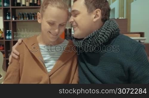 Happy young couple in cafe showing romantic feelings with hugs and kiss while making selfie using smart phone. Then they smiling and looking to camera