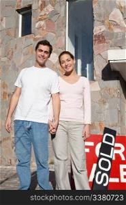 Happy young couple in front of their new home with sold sign in background