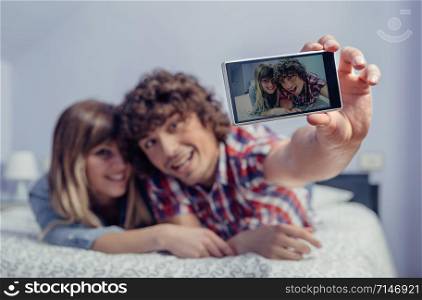 Happy young couple in love taking a selfie with smartphone lying over a bed. Selective focus on the phone.. Couple in love taking selfie with smartphone on bed