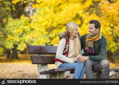 Happy young couple looking at each other while sitting on park bench during autumn