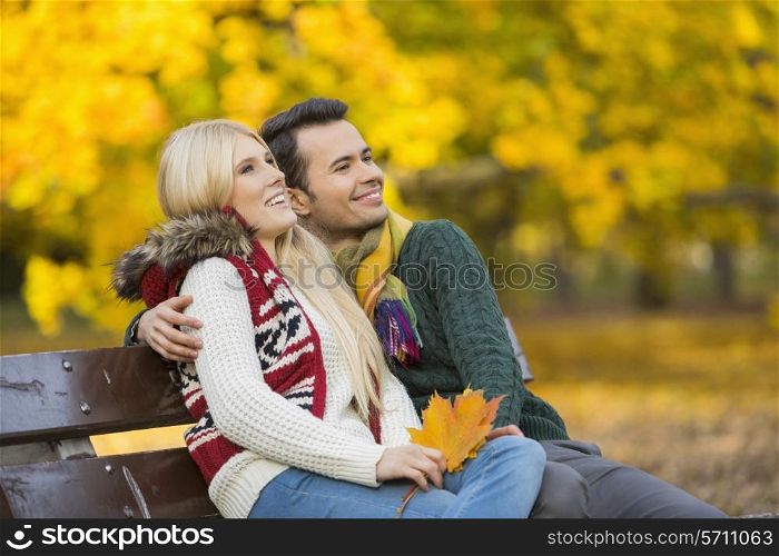 Happy young couple looking away while sitting on park bench during autumn