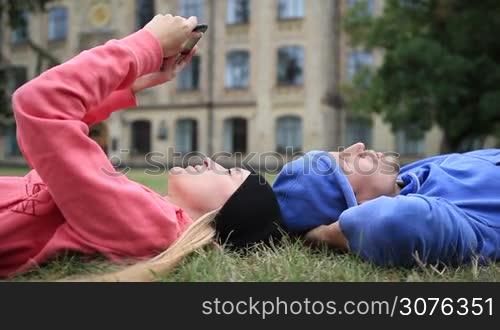 Happy young couple of students lying on the grass head to head and relaxing after studying. Smiling college students resting on campus lawn and talking while cute female student searching online on tablet. Side view camera.