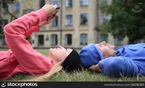 Happy young couple of students lying on the grass head to head and relaxing after studying. Smiling college students resting on campus lawn and talking while cute female student searching online on tablet. Side view camera.