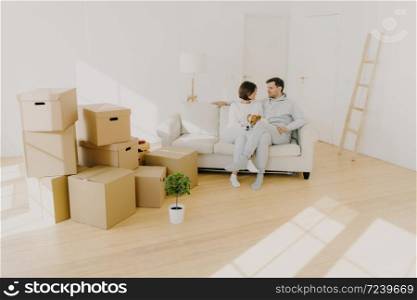 Happy young couple pose together on cozy sofa, have pleasant conversation, tease dog, relocate in new home, enjoy relax after unpacking personal belongings, stack of boxes near, house plant.