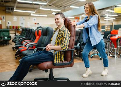 Happy young couple rides on office chair in furniture store showroom. Man and woman looking samples for bedroom in shop, husband and wife buys goods for modern home interior. Couple rides on office chair in furniture store