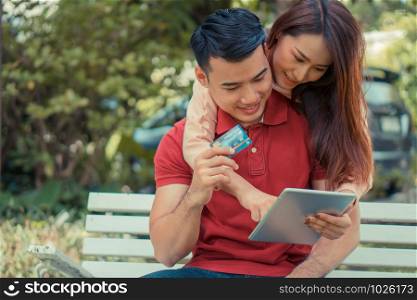 Happy young couple sitting in the garden and holding a tablet and dummy credit card and currently purchasing products via e-commerce. Concept of shopping on the internet with credit card payment.