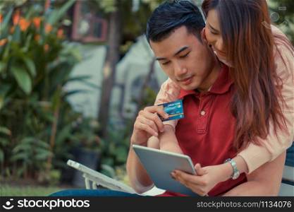 Happy young couple sitting in the garden and holding a tablet and dummy credit card and currently purchasing products via e-commerce. Concept of shopping on the internet with credit card payment.