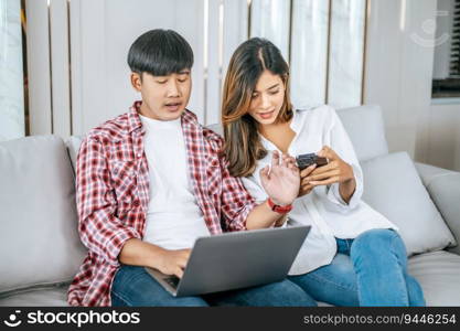 Happy Young couple sitting on sofa in living room at home use laptop computer working and smartphone, talking and smile together with happiness, happy family concept