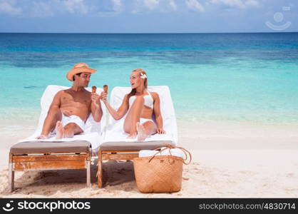 Happy young couple sitting on sunbed on the beach, eating tasty sweet cold ice cream, enjoying romantic summer vacation