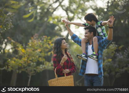 Happy young couple standing in a garden with the man carrying their son on his shoulders and the woman carrying a basket.