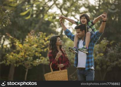 Happy young couple standing in a garden with the man carrying their son on his shoulders and the woman carrying a basket.