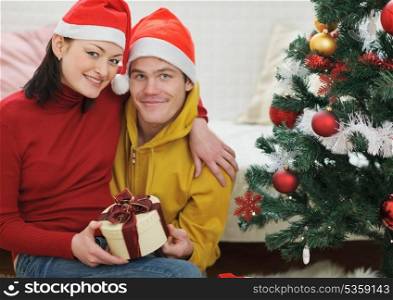 Happy young couple with gift sitting near Christmas tree