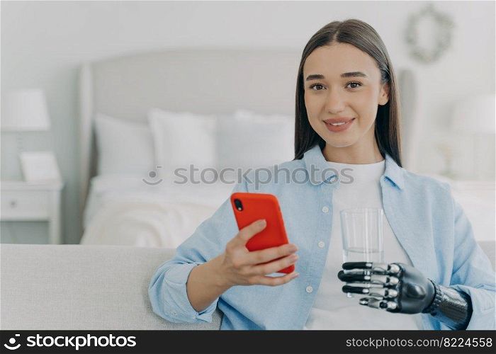 Happy young disabled girl holding mobile phone and glass of water using bionic prosthetic arm, sitting on couch at home. Female with disability uses apps on smartphone. Disability, healthy lifestyle.. Happy young disabled girl holding smartphone and glass of water, using bionic prosthetic arm