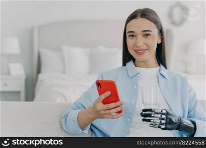 Happy young disabled girl holding mobile phone and glass of water using bionic prosthetic arm, sitting on couch at home. Female with disability uses apps on smartphone. Disability, healthy lifestyle.. Happy young disabled girl holding smartphone and glass of water, using bionic prosthetic arm