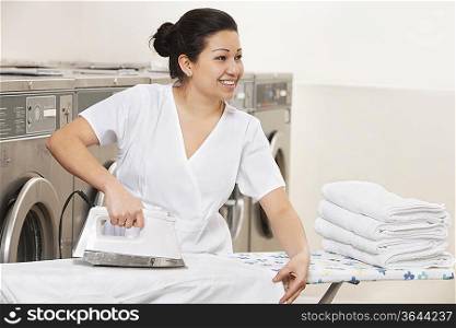 Happy young employee ironing clothes while looking away in Laundromat