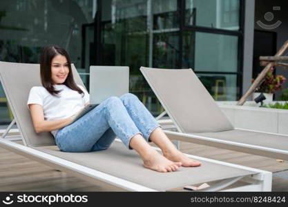 Happy young entrepre≠ur woman sitting on tanning bed beside pool and using laptop computer for remote onli≠working digital, onli≠busi≠ss project in quiet yard of resort house, Work on vacation
