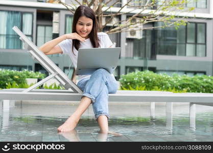 Happy young entrepreneur woman sitting on tanning bed beside pool and using laptop computer for remote online working digital, online business project in quiet yard of resort house, Work on vacation