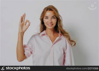 Happy young European female with fair hair with beaming smile showing ok sign while posing isolated against white studio wall background, smiling woman making okay sign with hand. Body language. Happy young European female showing ok sign with fingers while standing isolated on grey wall