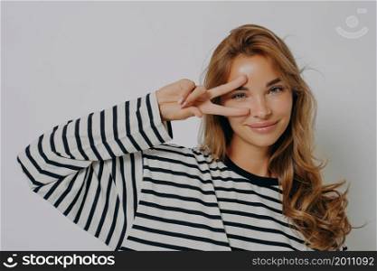 Happy young European woman makes peace disco gesture over eye v sign wears casual striped jumper minimal makeup stands indoor against grey studio background. People and body language concept. Happy young European woman makes peace disco gesture over eye v sign wears casual striped jumper