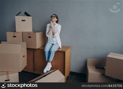 Happy young european woman purchases real estate and thanks god. Girl is sitting on boxes in new apartment and dreaming. Mover is excited. Concept of success and opportunities for women.. Happy young european woman purchases real estate. Thanks god. Girl is sitting on boxes and dreaming.