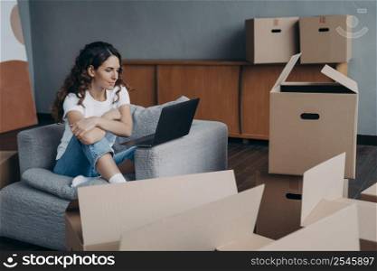 Happy young european woman selling apartment through internet. Girl among cardboard boxes is using laptop and smiling. Lady is packing things. Relocation concept.. Happy young european woman selling apartment through internet. Girl among boxes is using laptop.