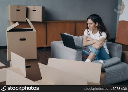 Happy young european woman selling apartment through internet. Girl among cardboard boxes is using laptop and smiling. Lady is packing things. Relocation concept.. Happy young european woman selling apartment through internet. Girl among boxes is using laptop.