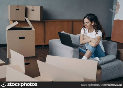 Happy young european woman selling apartment through internet. Girl among cardboard boxes is using laptop and smiling. Lady is packing things. Relocation concept.. Happy young european woman selling apartment through internet. Girl among boxes is using laptop.