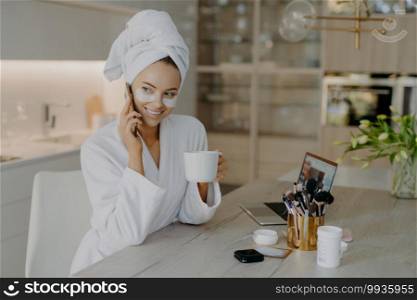 Happy young European woman undergoes beauty procedures after morning shower wears hydrogel moisturising patches dressed in white bathrobe drinks aromatic coffee talks via mobile phone poses indoor
