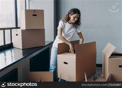 Happy young european woman unpacking boxes. Girl near window in white t-shirt indoors. Woman is opening cardboard box and smiling. Relocation and independence concept.. Happy young european woman unpacking boxes. Girl near window opening cardboard box and smiling.