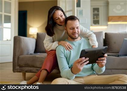 Happy young family couple using mobile tablet at home together. Positive smiling wife and husband surfing on Internet, doing online shopping or booking ticket, talking friend via web chat. Happy young family couple using mobile tablet at home together