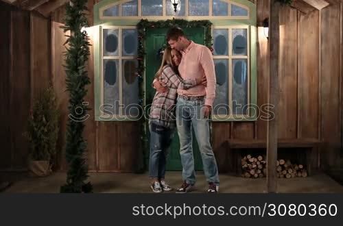 Happy young family with cute daughter embracing and smiling while standing on the porch of Christmas decorated house in the evening. Father holding adorable girl in his arms and hugging beloved wife. Family looking at camera and smiling.